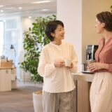 職場のコーヒースペースで外国人の同僚と英語で挨拶する大人女性
